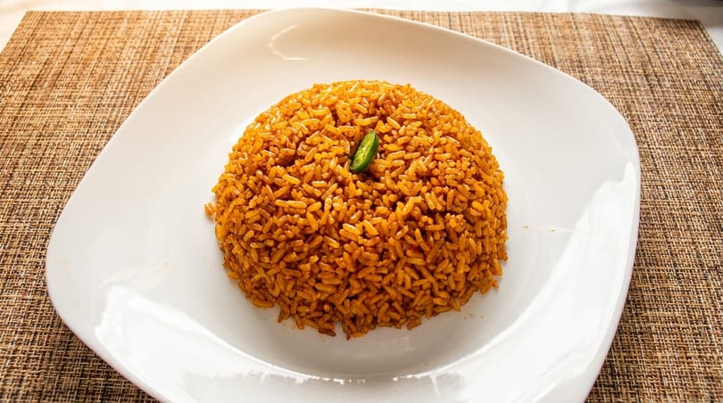 A mound of orange-red jollof rice with a green chili pepper on top, served on a white plate.