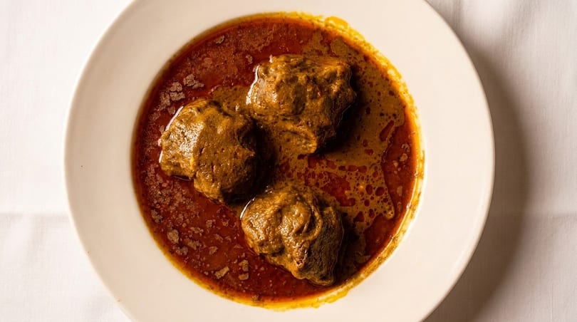 Three large meat pieces in a rich, reddish-brown curry sauce, served in a white bowl.